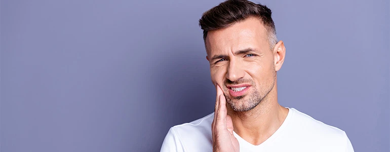 Zu sehen ist ein Mann, der Kieferschmerzen hat und sich seine Hand seitlich an sein Gesicht legt; sein Gesichtausdruck ist leicht schmerzverzerrt.